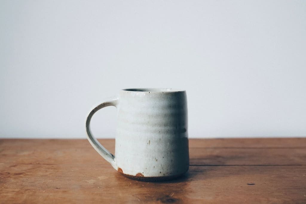 Tazza grigio chiaro con manico, su un tavolo di legno davanti a un muro bianco.