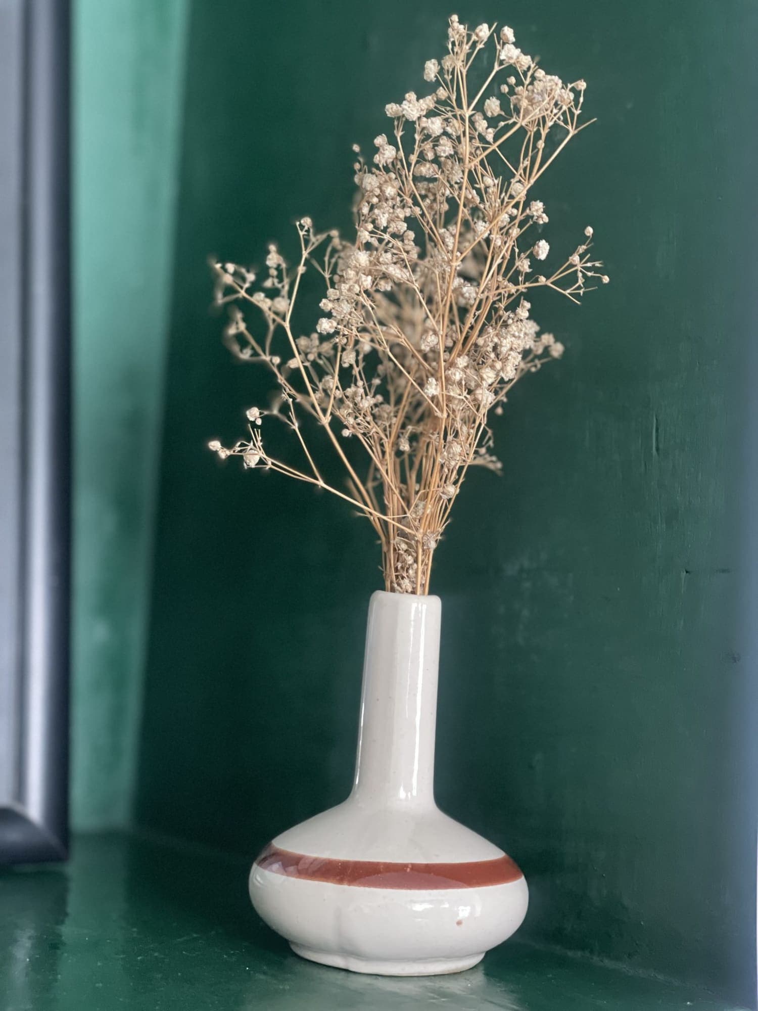 Bottiglia bianca stretta con fiori di campo secchi davanti a un muro verde.